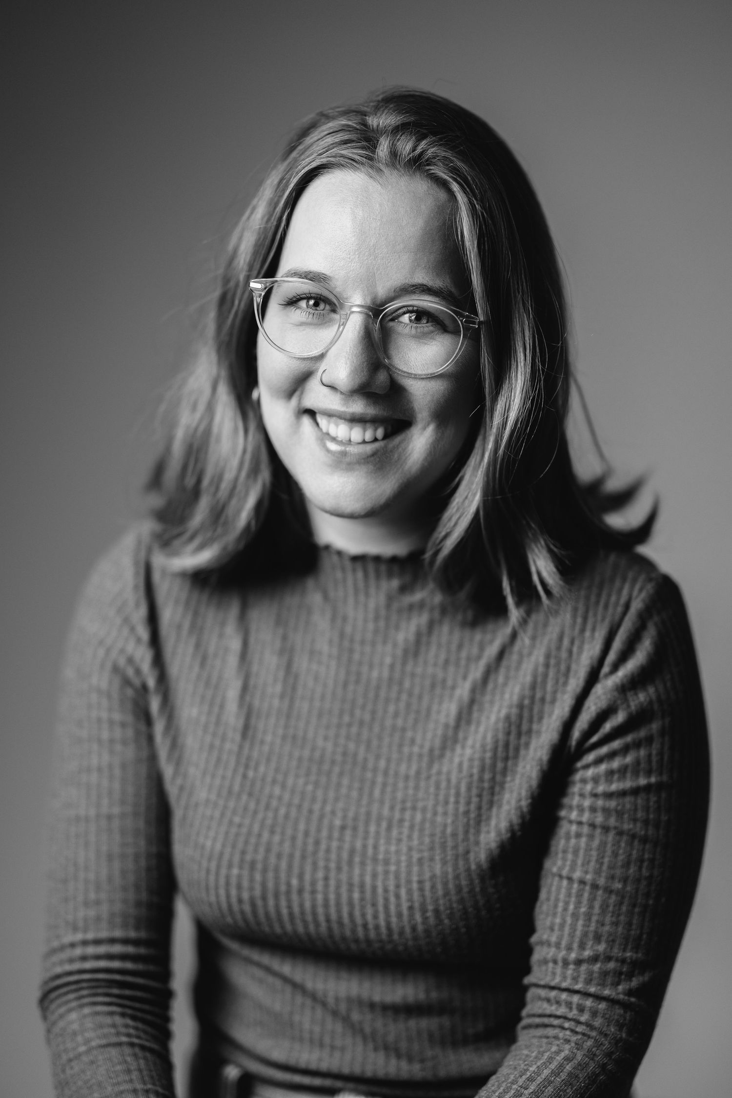 Black and white headshot of a woman in a ribbed roll-neck, wearing glasses, smiling, grey background