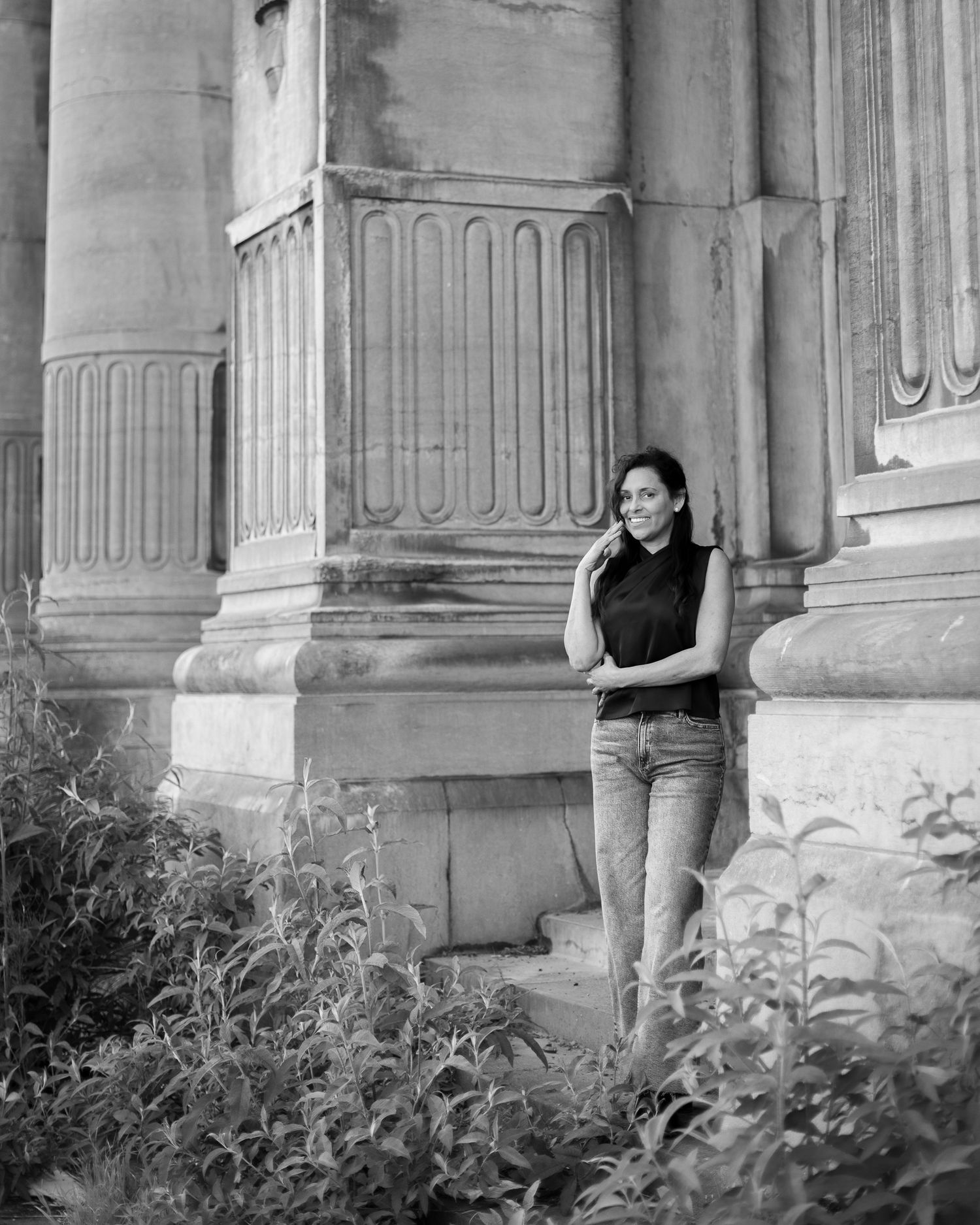 Black and white portrait of a woman in a sleeveless top standing beside tall stone columns, wild plants at the base