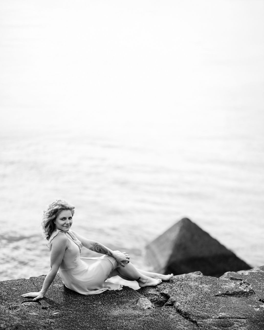 Black and white portrait of a woman in a light sundress sitting on coastal rocks by calm water, smiling