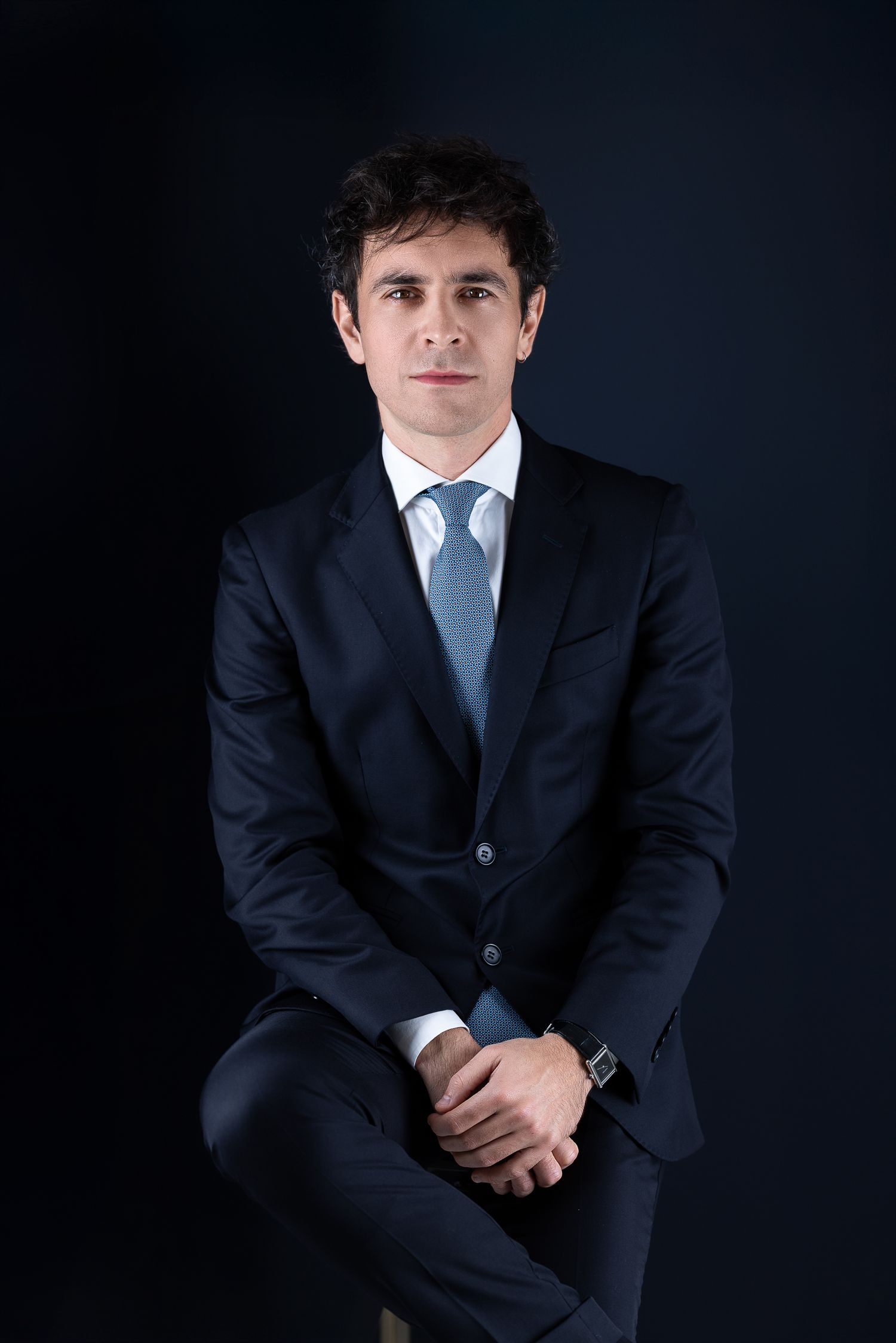 Professional portrait of a young man in a dark navy suit with a light blue tie, seated, hands clasped