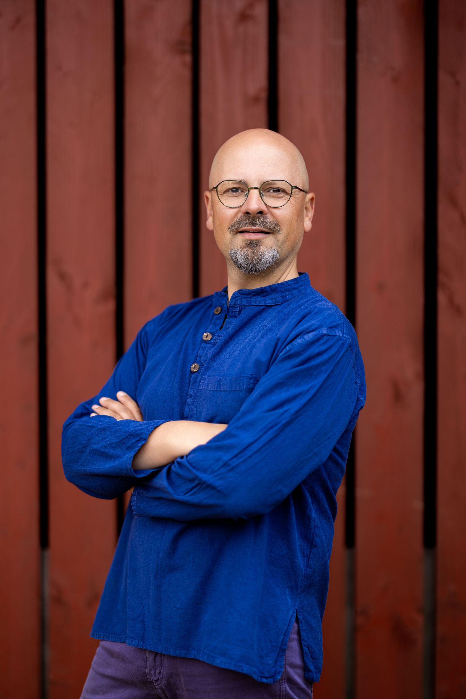 Colour portrait of a bald man with a beard and glasses in a bright blue shirt, arms crossed, red timber panel backdrop