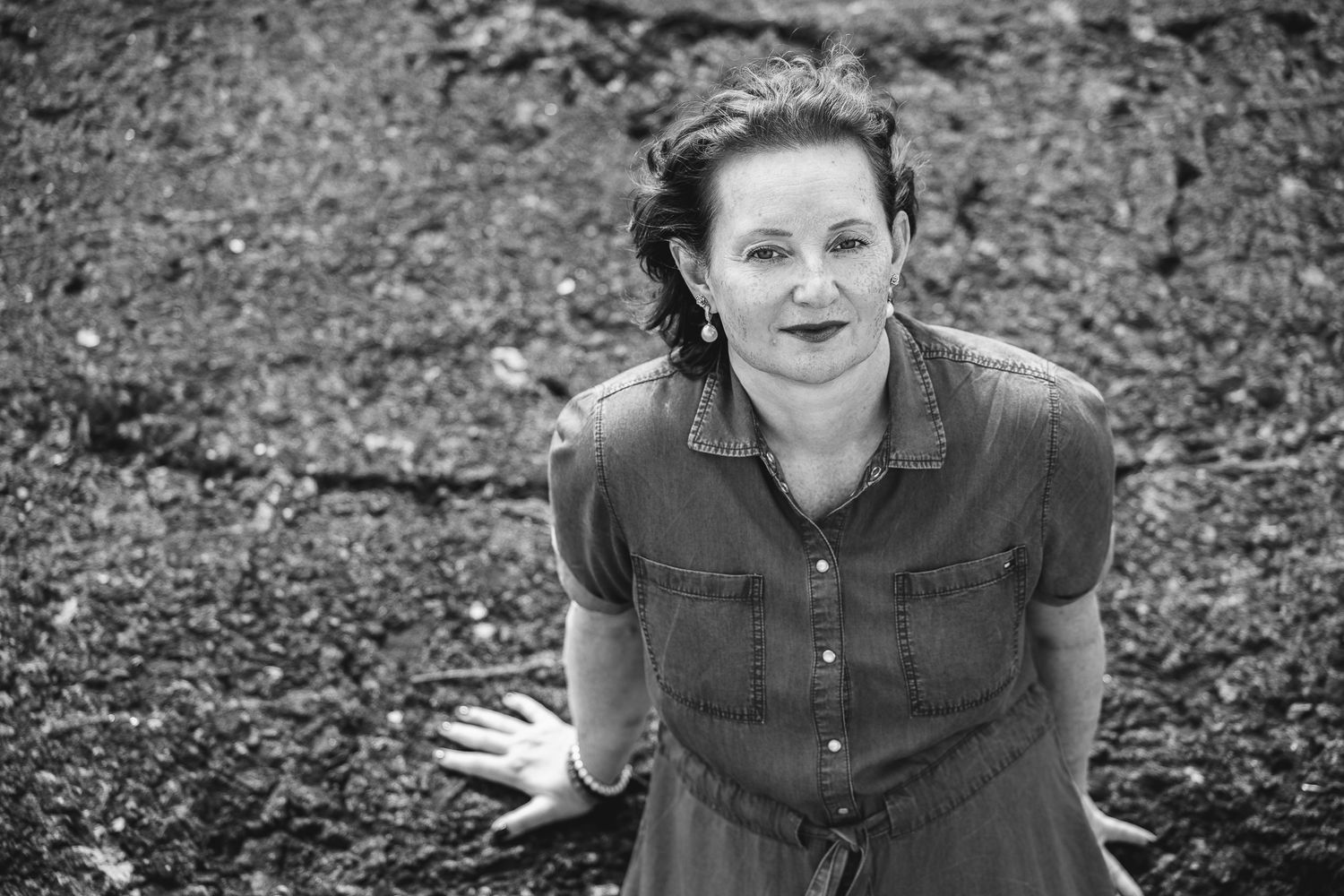 Black and white portrait of a woman in a denim dress leaning forward with hands spread, rocky backdrop