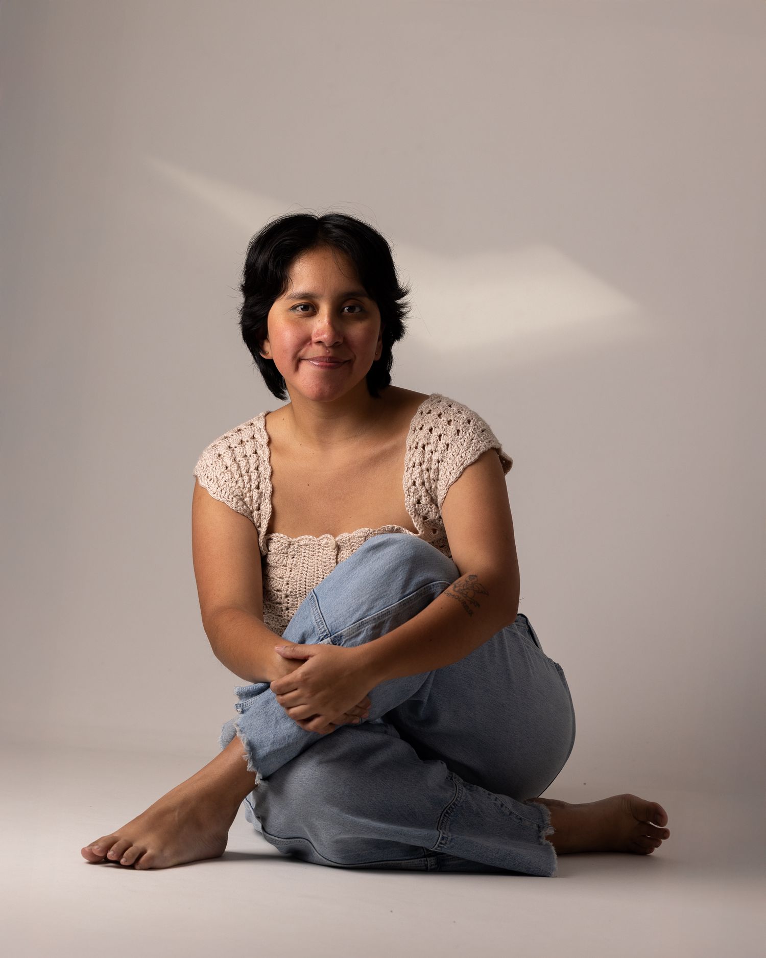 Colour portrait of a woman in a crochet top and jeans, seated cross-legged on the floor, smiling