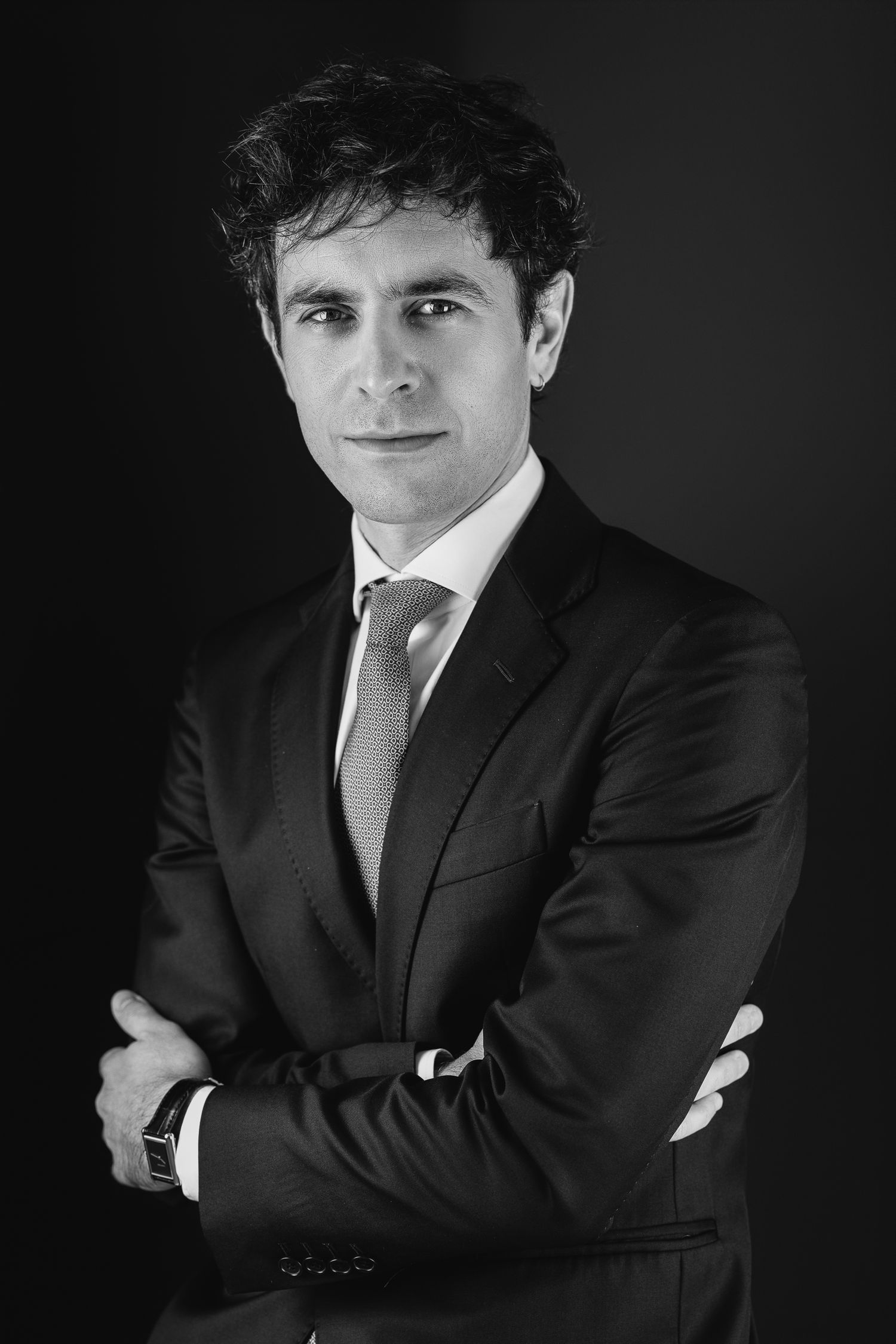 Black and white professional headshot of a man in a dark suit and tie, arms crossed, dark background