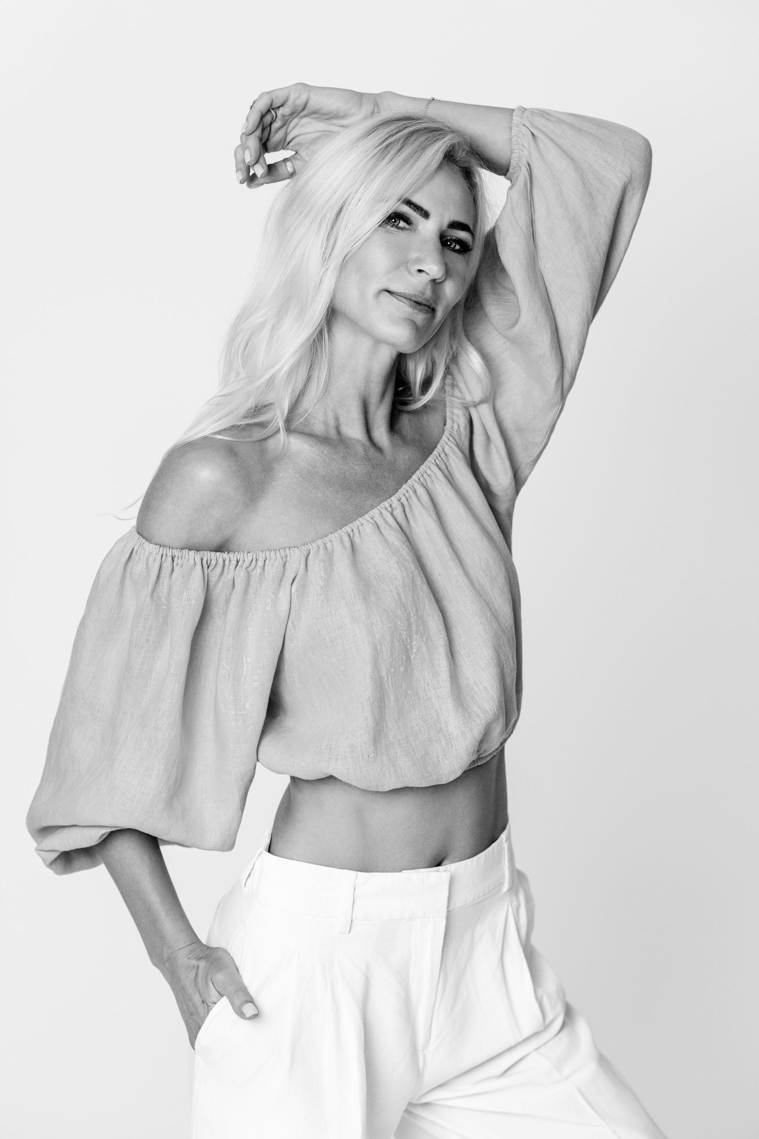 Black and white portrait of a woman in an off-shoulder top, arm raised, in an airy outdoor setting