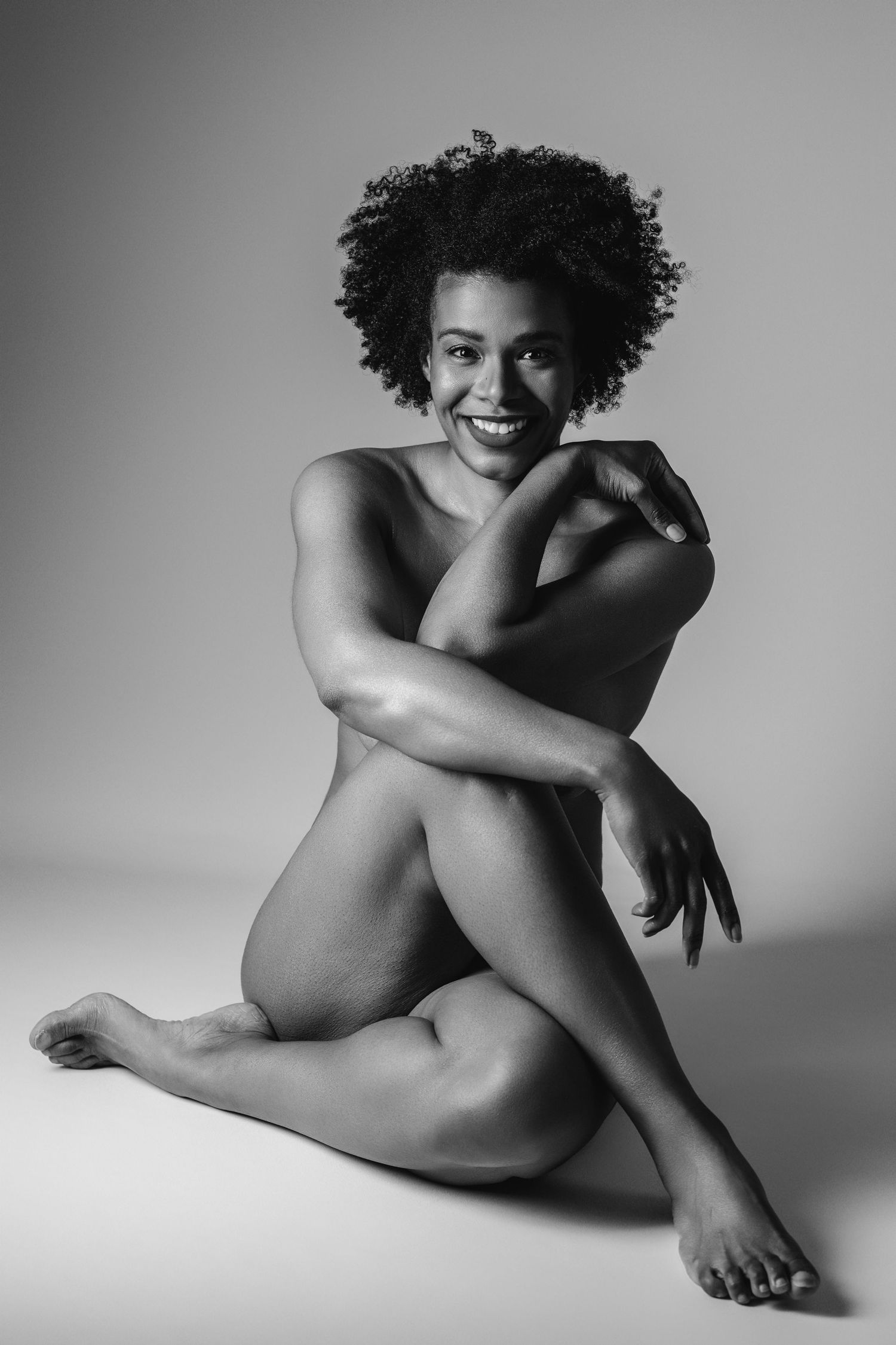 Black and white boudoir portrait of a woman with natural hair seated on a studio floor, arms crossed, smiling