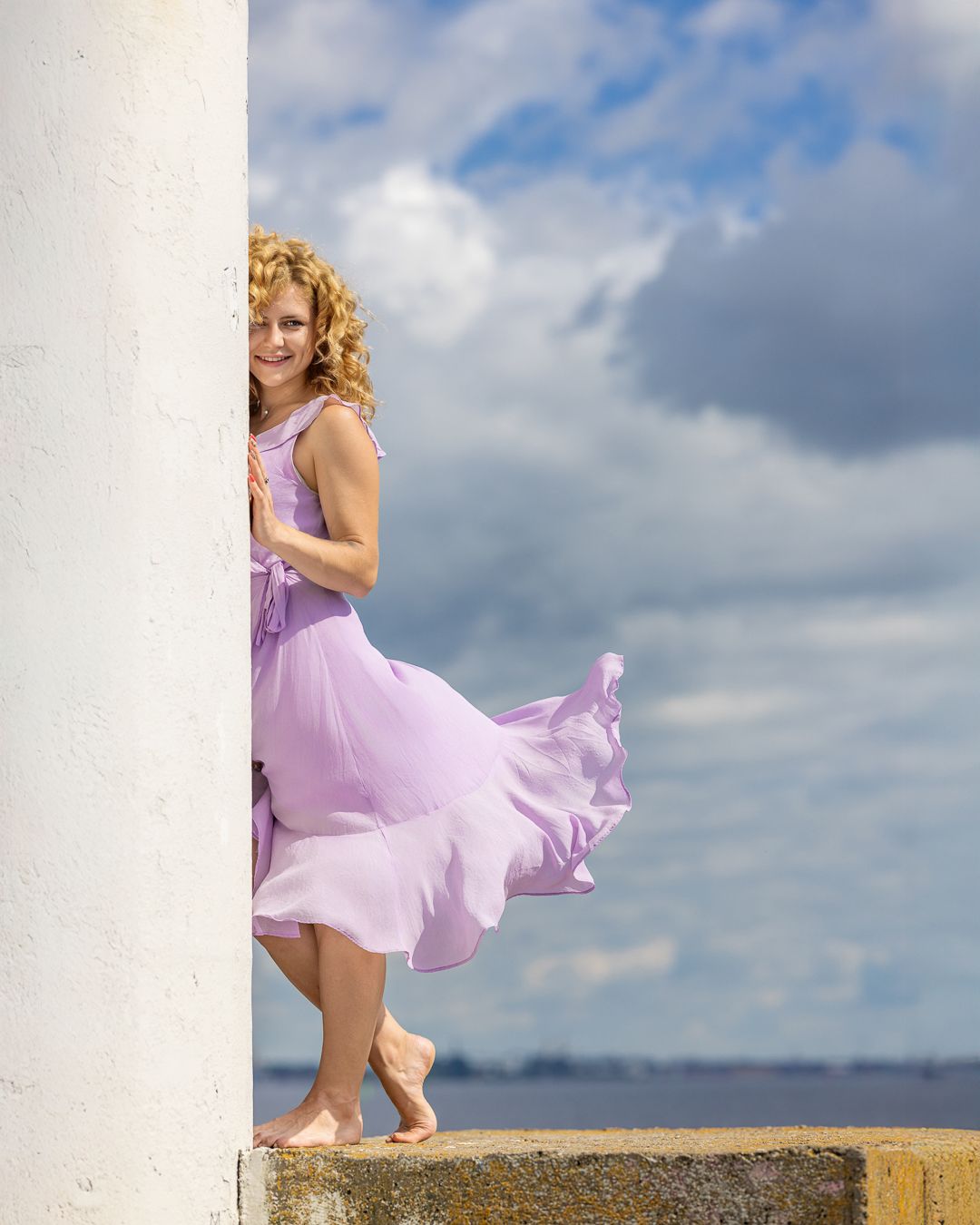 Colour portrait of a woman with curly hair in a lilac dress beside a white column, cloudy sky behind, dress blowing in the wind