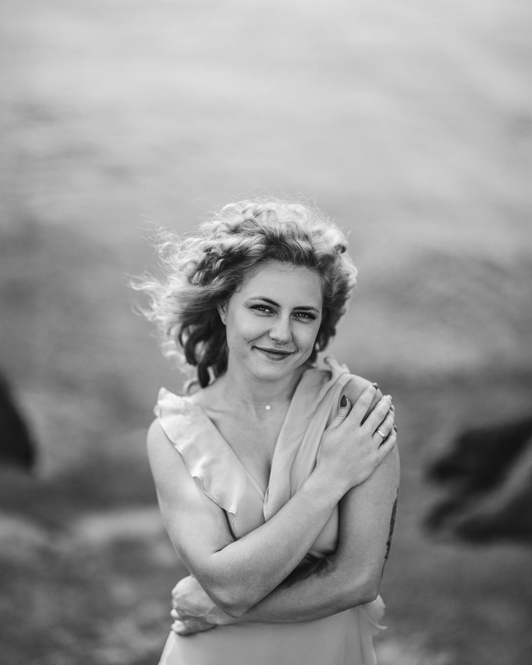 Black and white portrait of a woman with curly hair, arms crossed, smiling, outdoors against soft sky