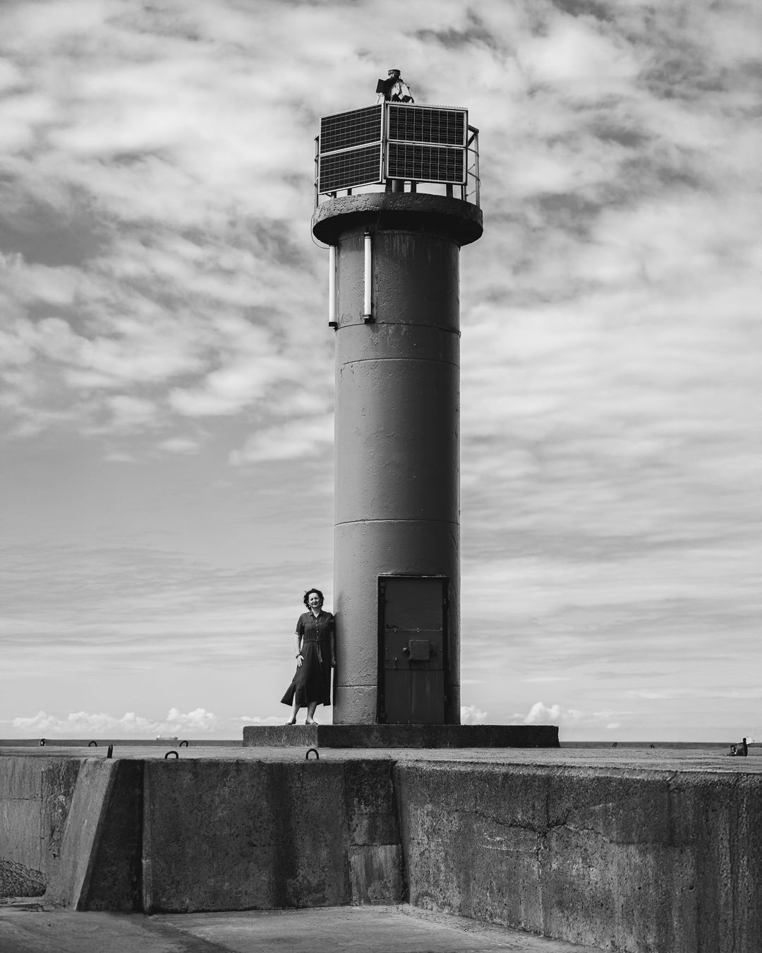 Black and white portrait of a woman in a dark dress standing at the base of a harbour lighthouse on a concrete pier