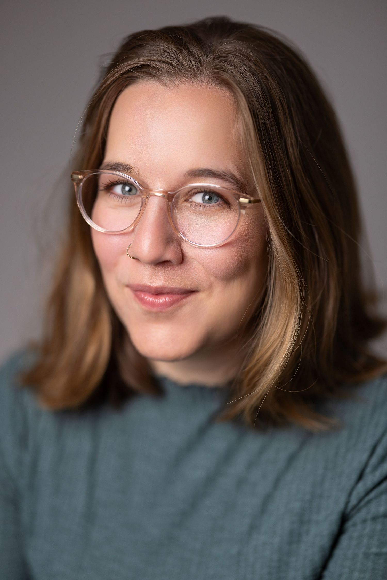 Colour headshot of a woman with light brown hair and round glasses in a teal top, grey background