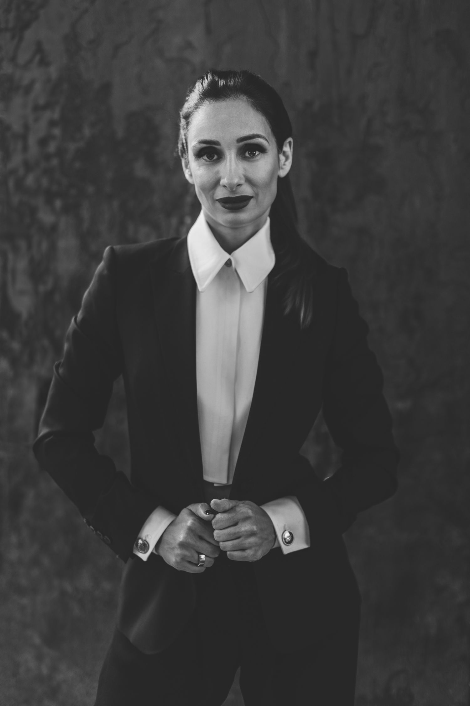 Black and white portrait of a woman in a dark suit jacket and white shirt, hands clasped, textured dark background