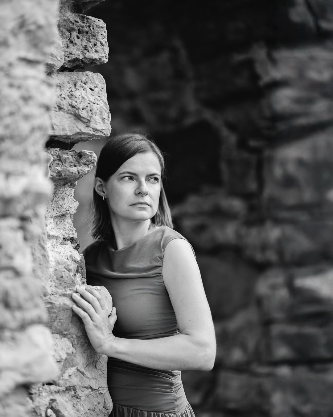 Black and white portrait of a woman in a dark sleeveless top leaning against a stone ruin wall, arms crossed