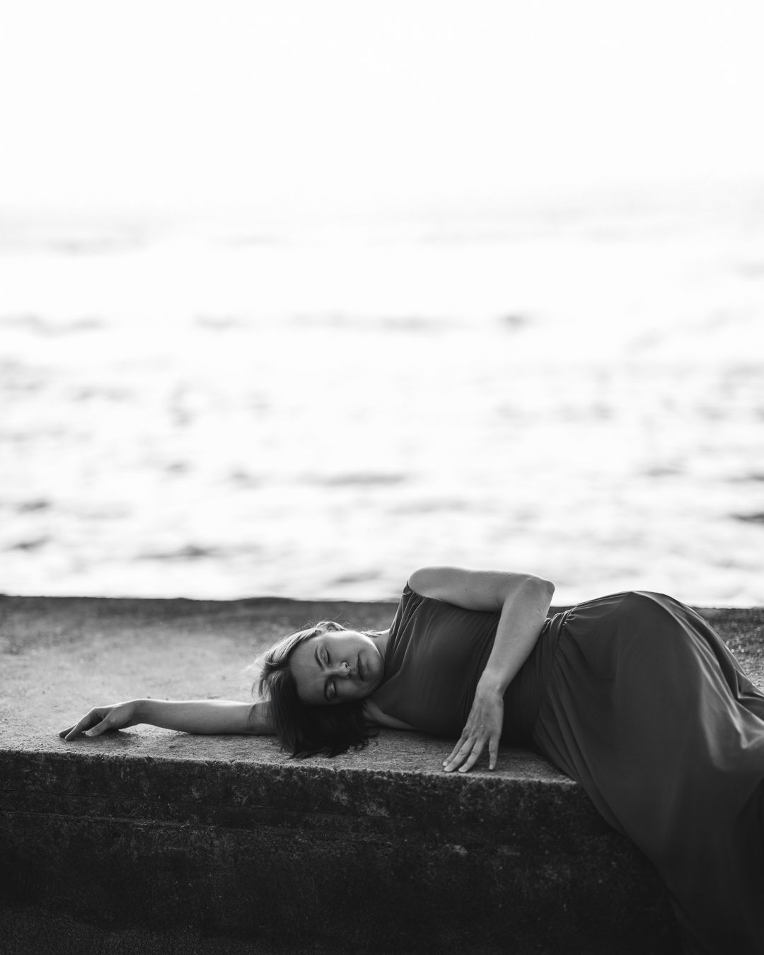 Black and white portrait of a woman in a dark dress reclining on a stone ledge beside the sea