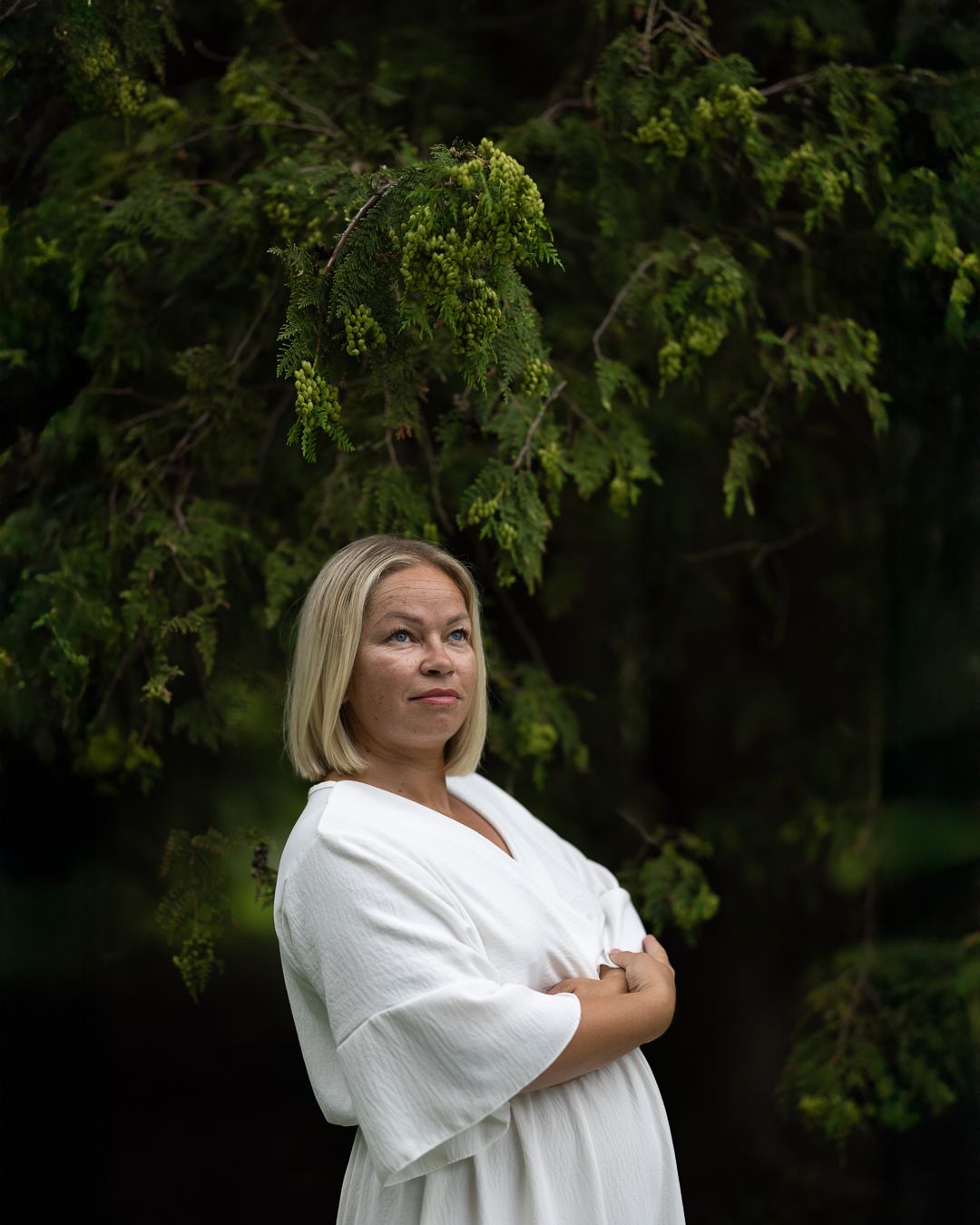 Colour portrait of a woman with blonde hair in a white blazer, arms crossed, dark green trees behind