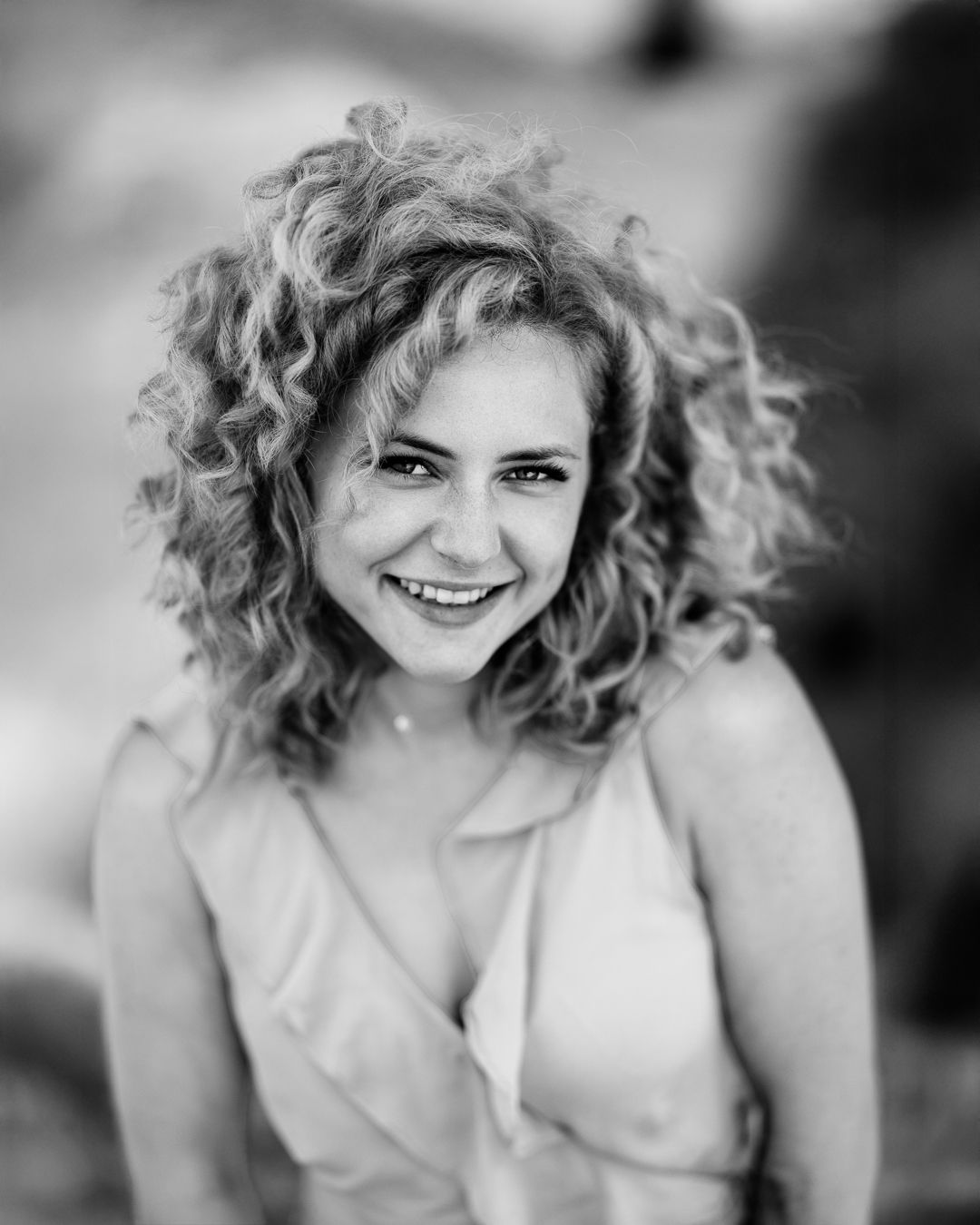 Black and white close-up portrait of a woman with curly hair, smiling, with a soft-focus outdoor background
