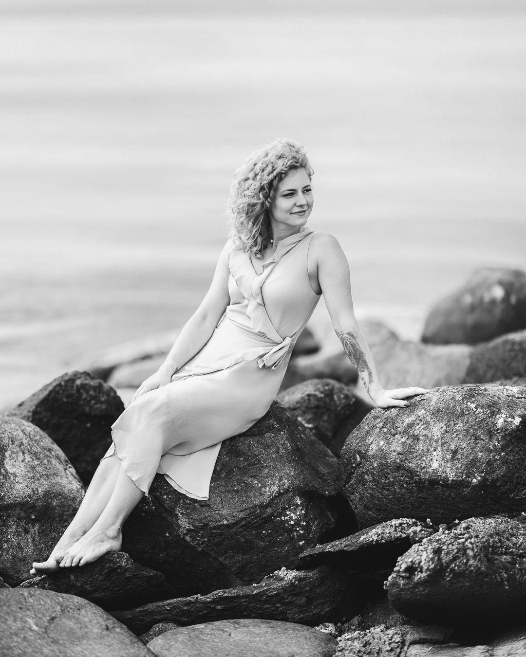 Black and white portrait of a woman in a light dress sitting on coastal rocks beside the sea, relaxed pose
