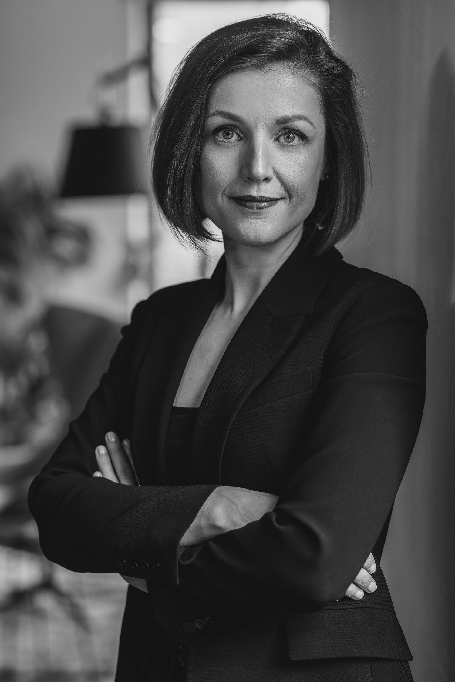 Black and white portrait of a woman in a dark blazer, arms crossed, with a soft interior background