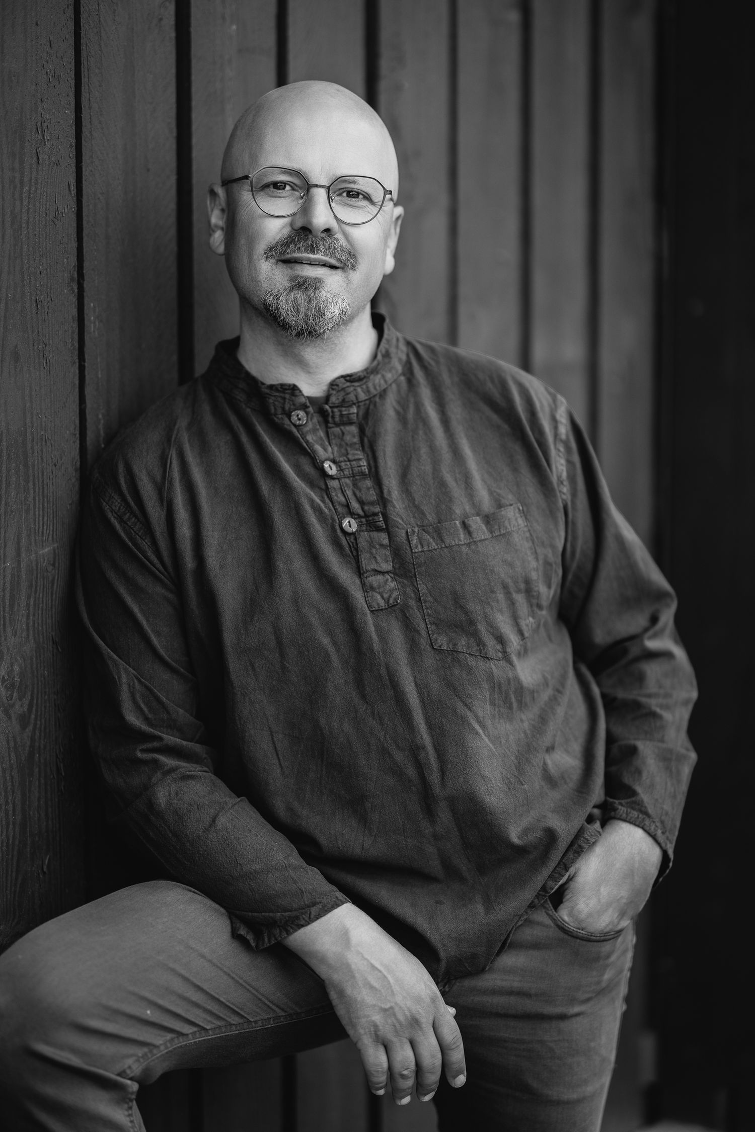 Black and white portrait of a bald man with glasses in a dark casual shirt, seated against a wooden panel background