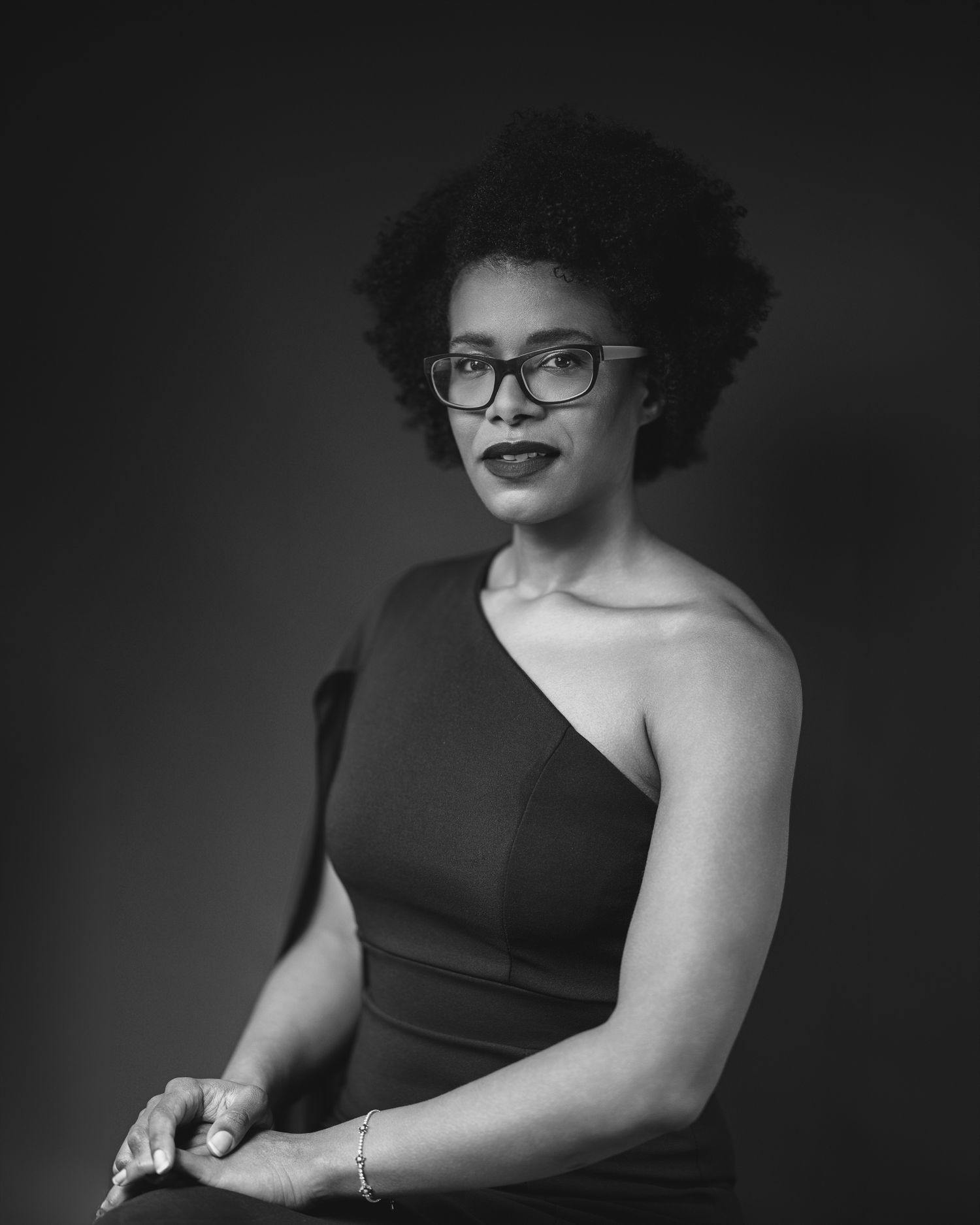 Black and white studio portrait of a woman with natural hair and glasses in a one-shoulder top
