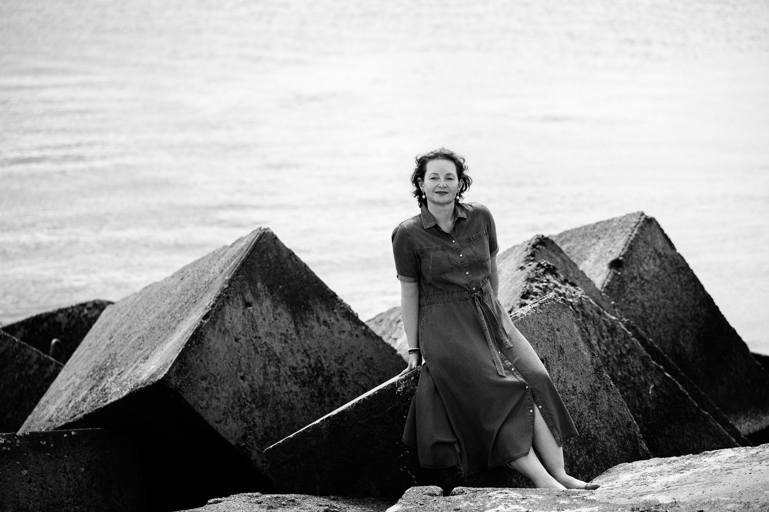 Black and white portrait of a woman in a midi dress leaning against large concrete sea-defence blocks by the water