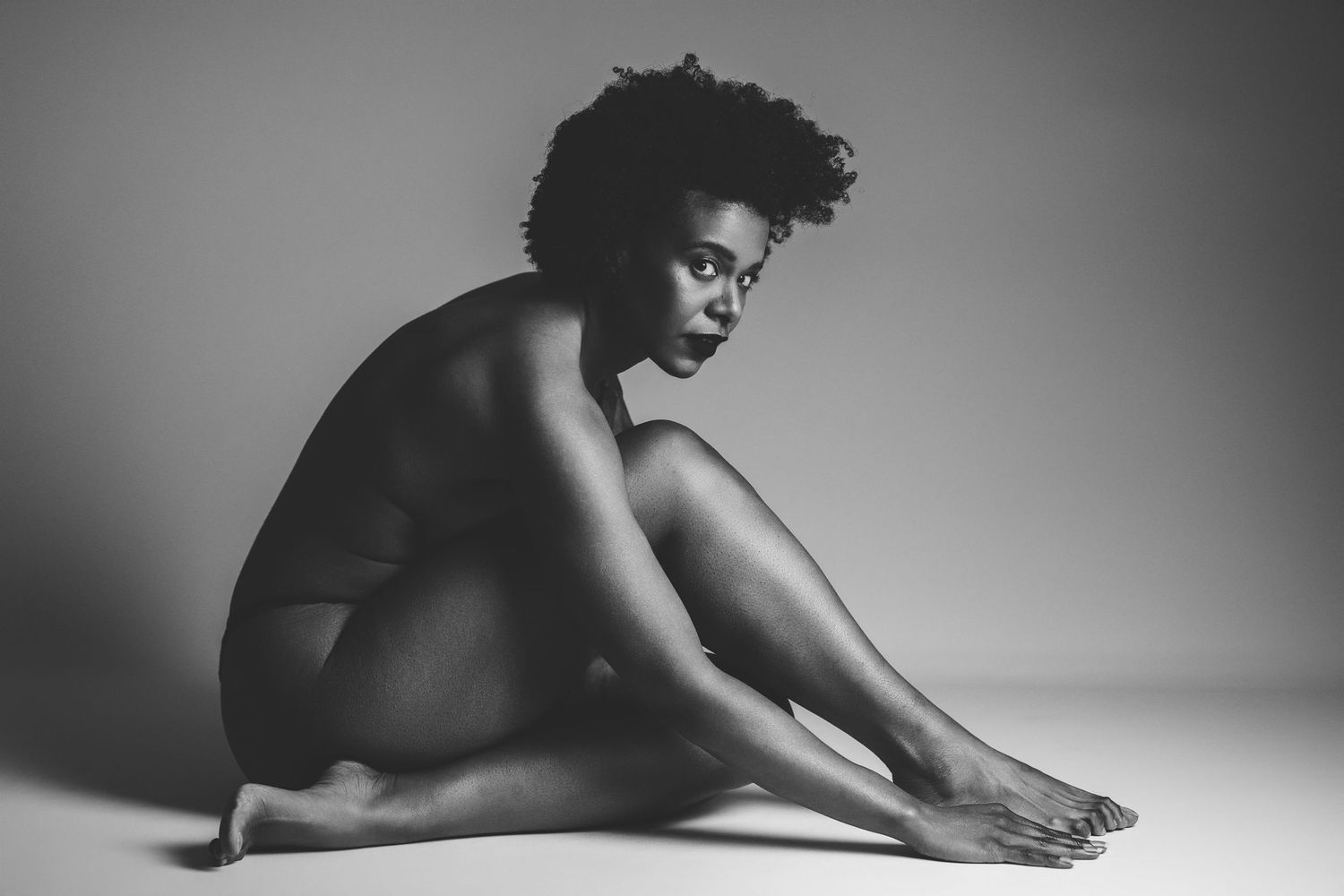 Black and white boudoir portrait of a woman with natural hair seated on a studio floor, composed pose in soft studio light