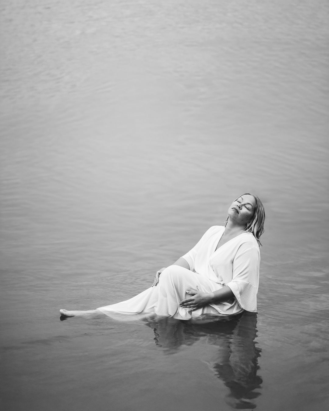 Black and white portrait of a woman in a white robe seated in still open water, head tilted back