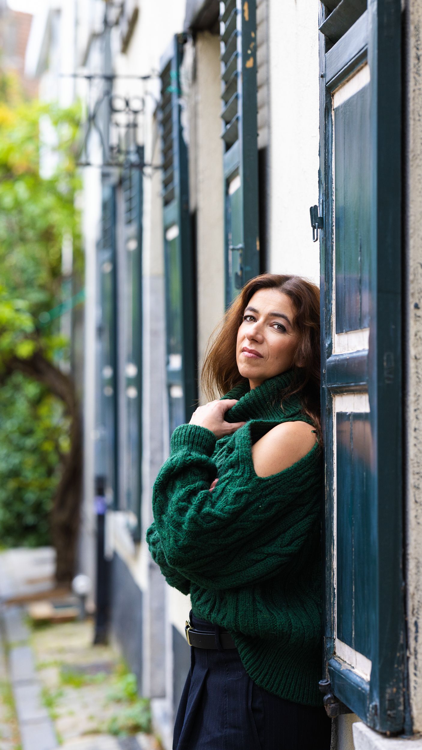 Colour portrait of a woman in a green jumper standing in a Brussels street beside blue shuttered windows