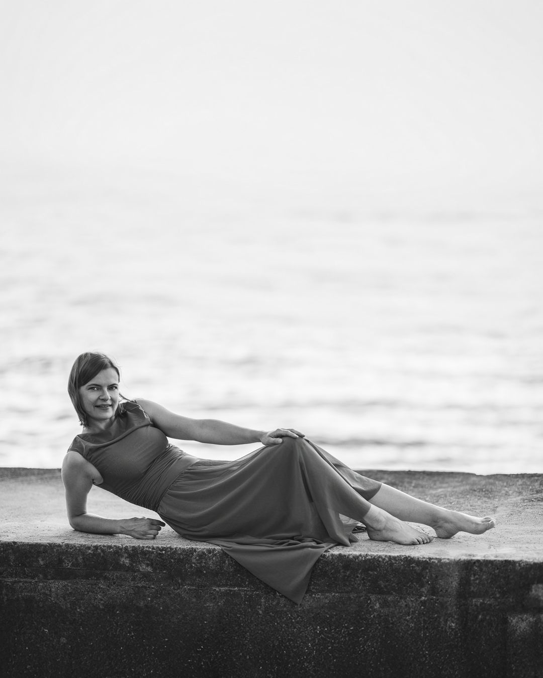 Black and white portrait of a woman in a midi dress reclining on a stone ledge beside open water