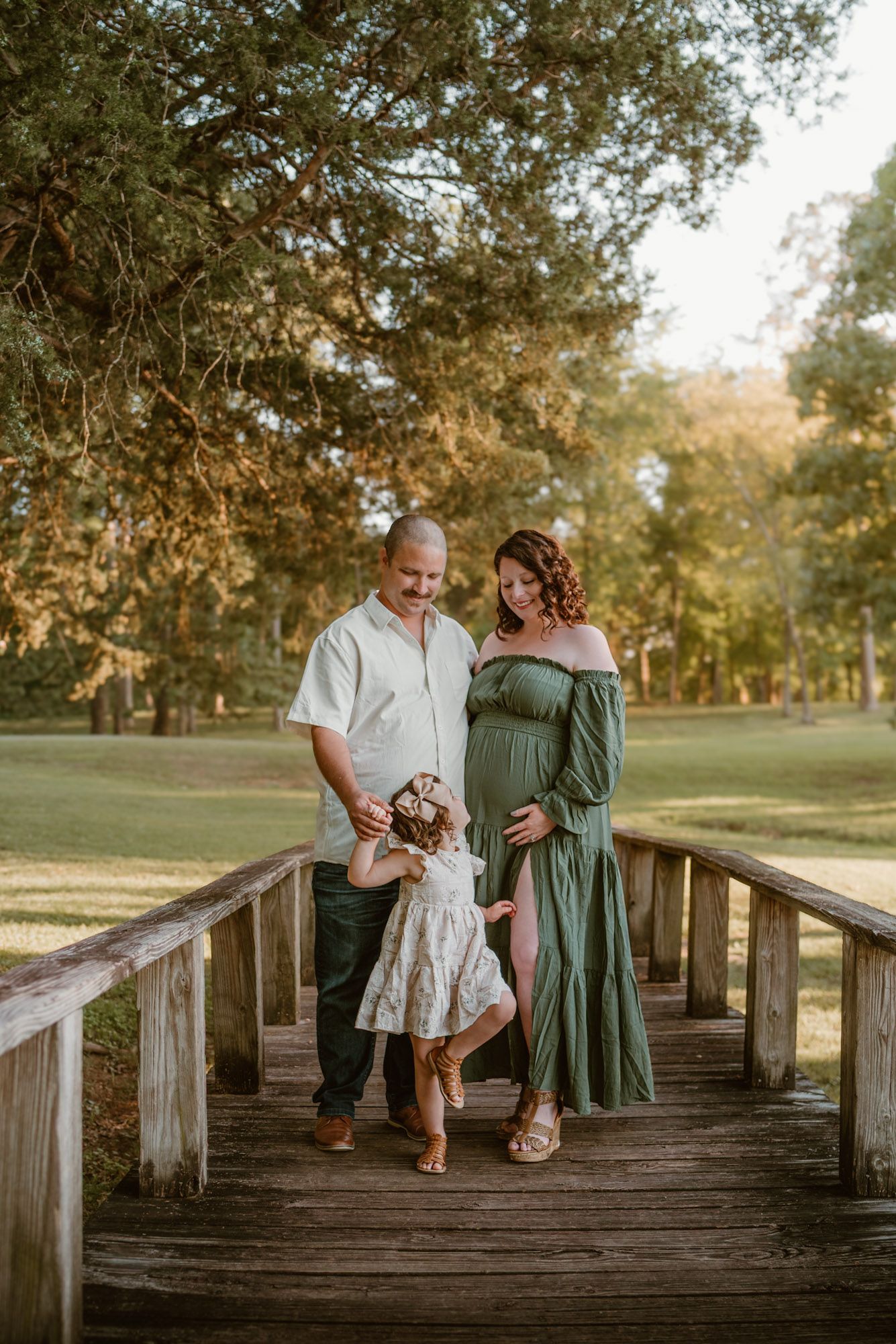 expecting couple standing on bridge smiling down at toddler in golden hour maternity session in Hixson TN