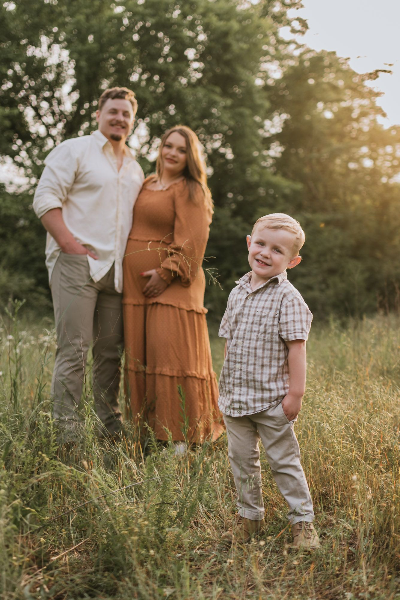 Expecting couple in background with toddler standing smiling at camera in field, family maternity session raccoon mountain Chattanooga TN