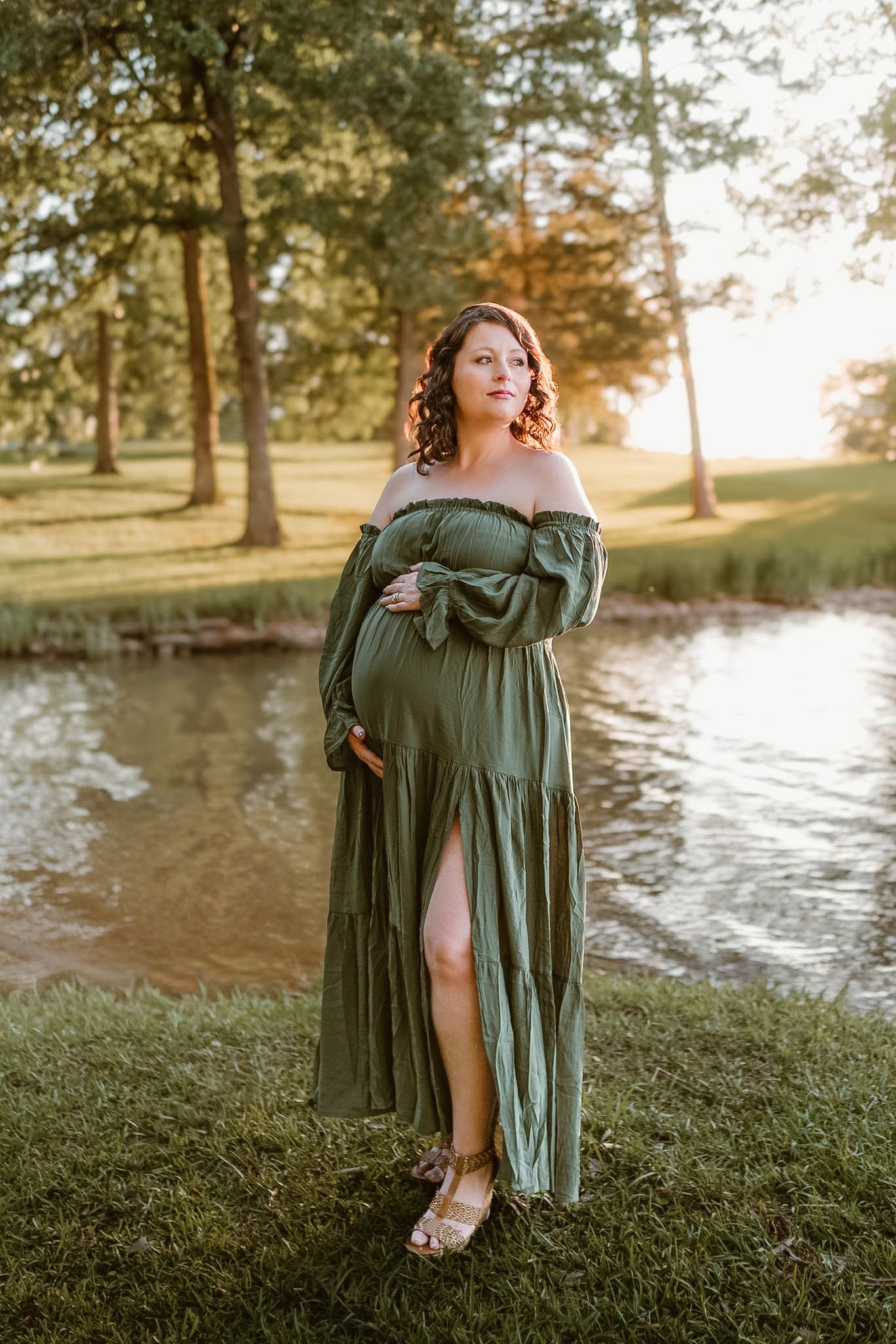 Maternity full body shot of mom holding belly and dress, looking off her shoulder in Hixson TN, warm golden light, bay in the background