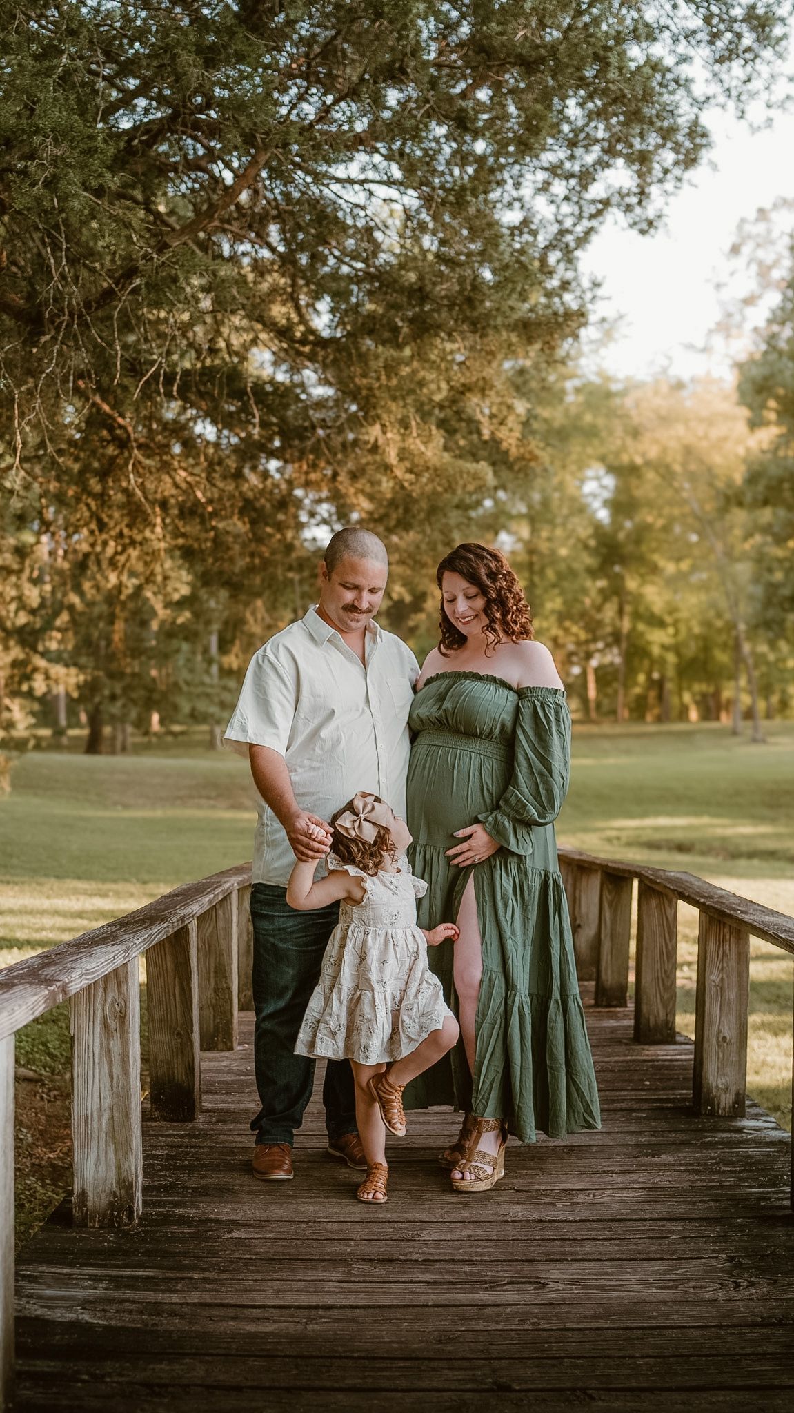 Posed family photo, with parents and toddler looking at each other on a bridge in a field in Hixson, Tennessee, golden hour light