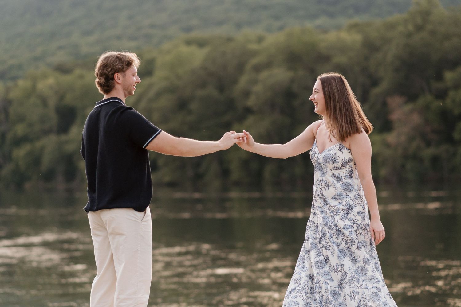 Couple holding hands far apart looking at each other with river and raccoon mountain behind them