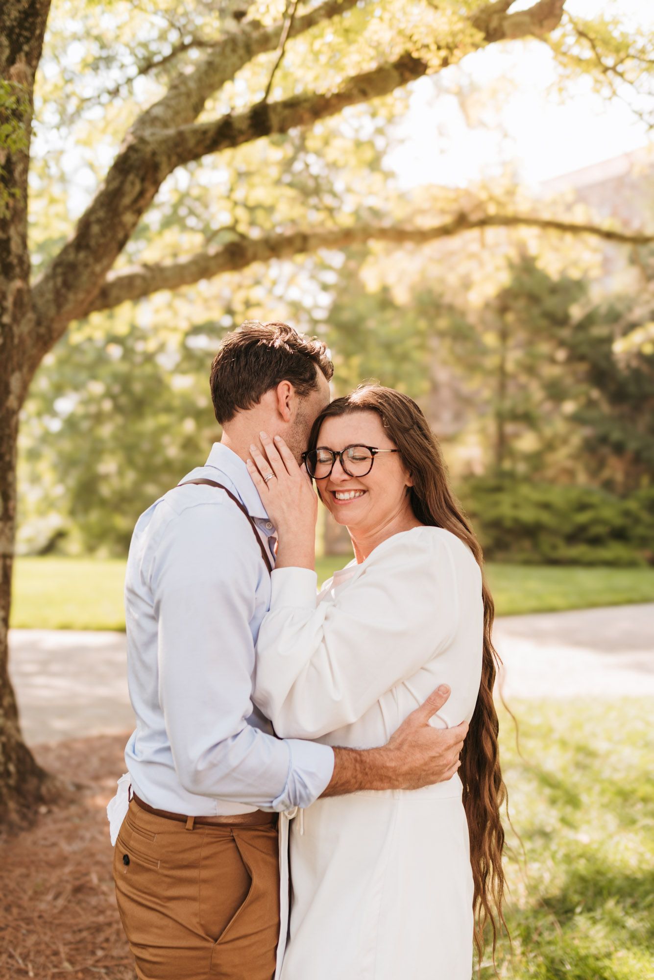 Couple standing close cheeks touching while woman laughs eyes closed, surrounded by soft natural light and trees on lookout mountain, GA