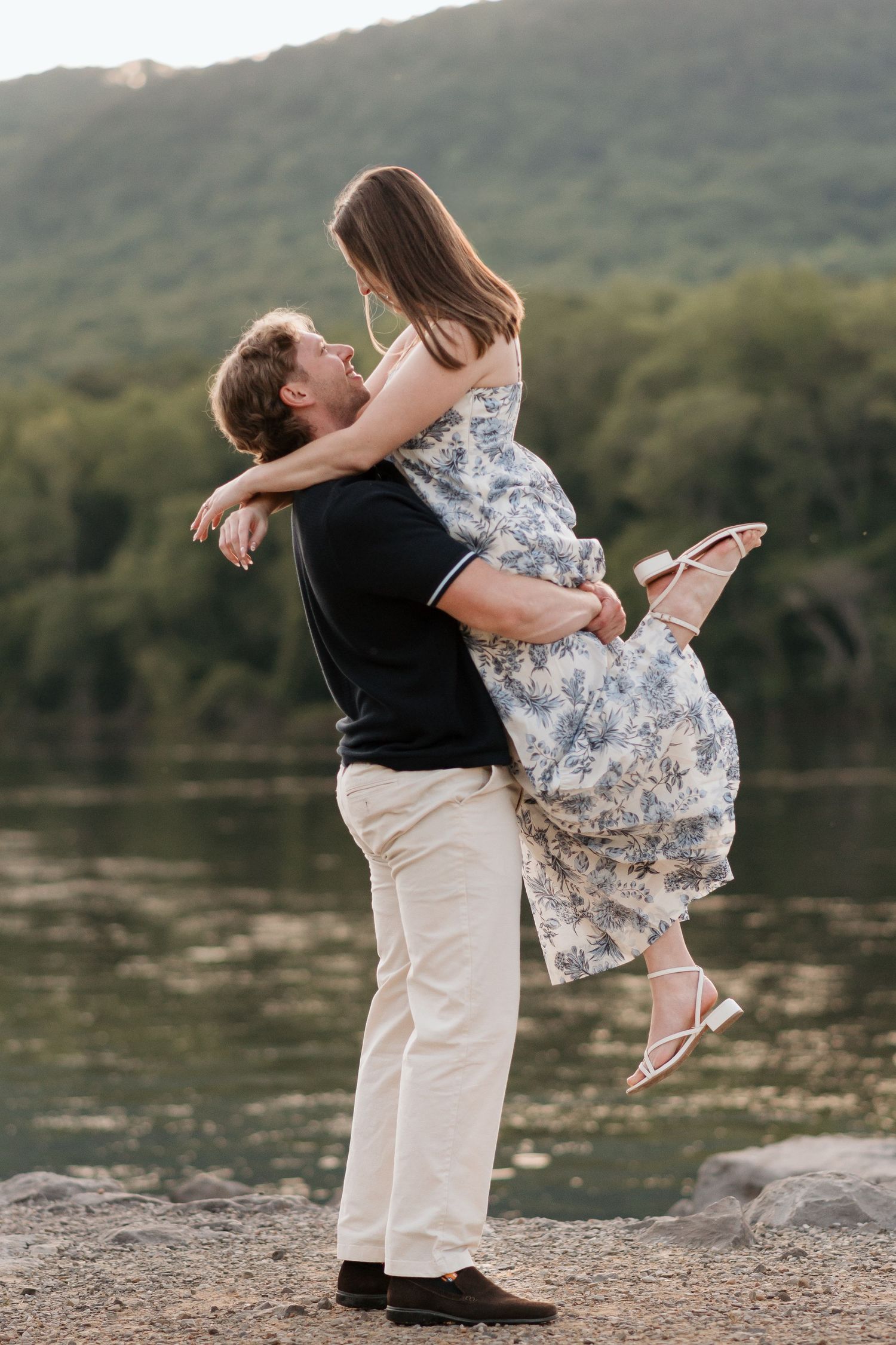 man holding fiance up as she kicks her legs out and holds her arms around his neck, looking at each other near river and raccoon mountain