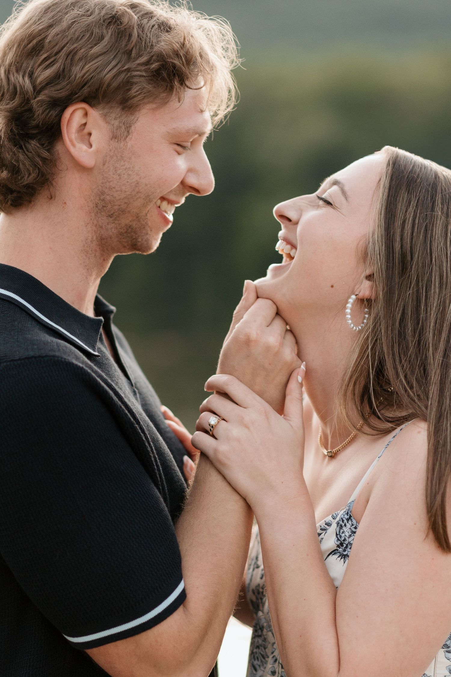 Close up of couple looking and laughing towards each other, holding her chin wile she holds his wrist, close up of engagement ring