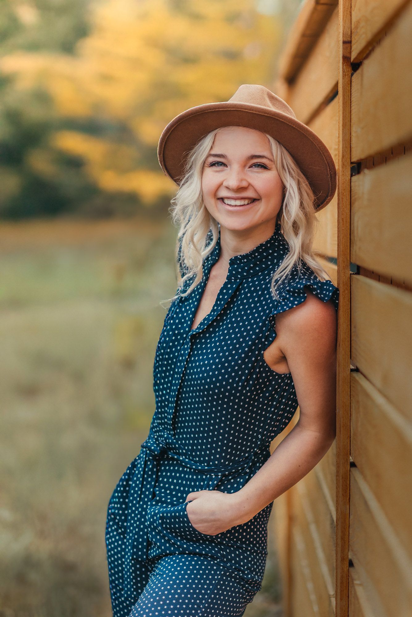 Chattanooga lifestyle photographer outdoors leaning on fence smiling at camera, wearing a blue jumpsuit and wide brim hat