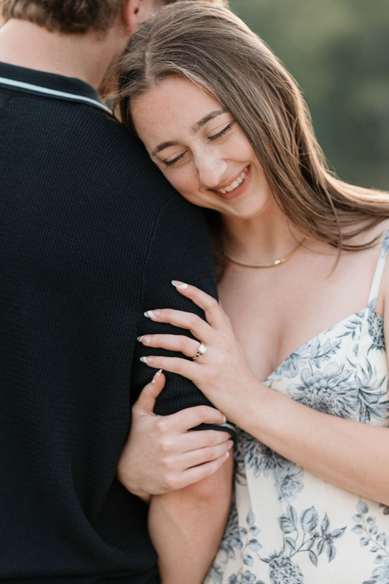 Woman smiling and looking down while holding and resting her head on mans arm, close up of engagement ring during proposal session