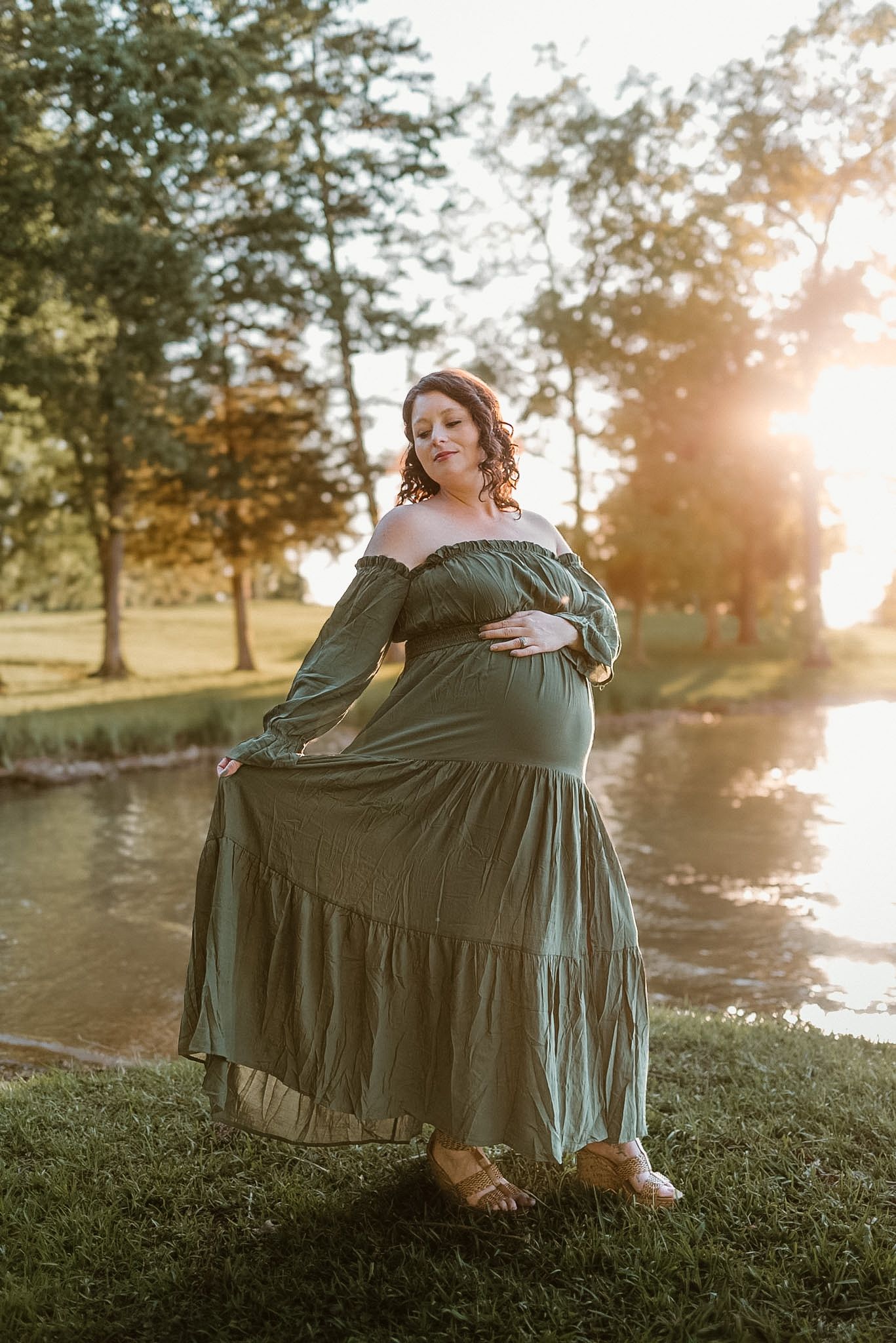 Maternity full body shot of mom holding belly and dress, looking off her shoulder in Hixson TN, warm golden light, bay in the background