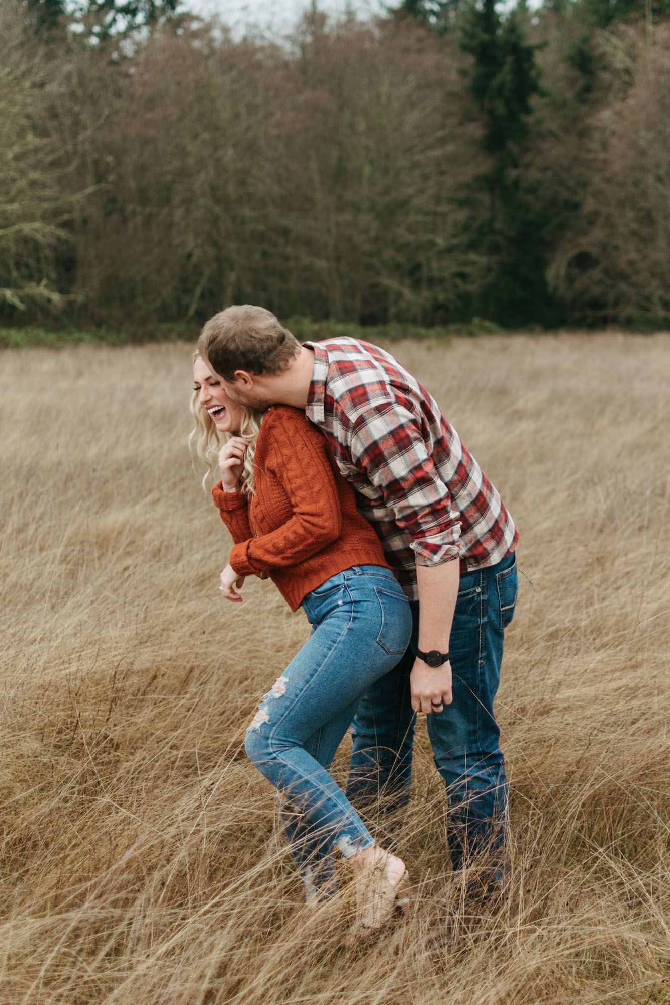 Playful portrait of couple standing close, him kissing her cheek from behind during outdoor fall couples engagement session