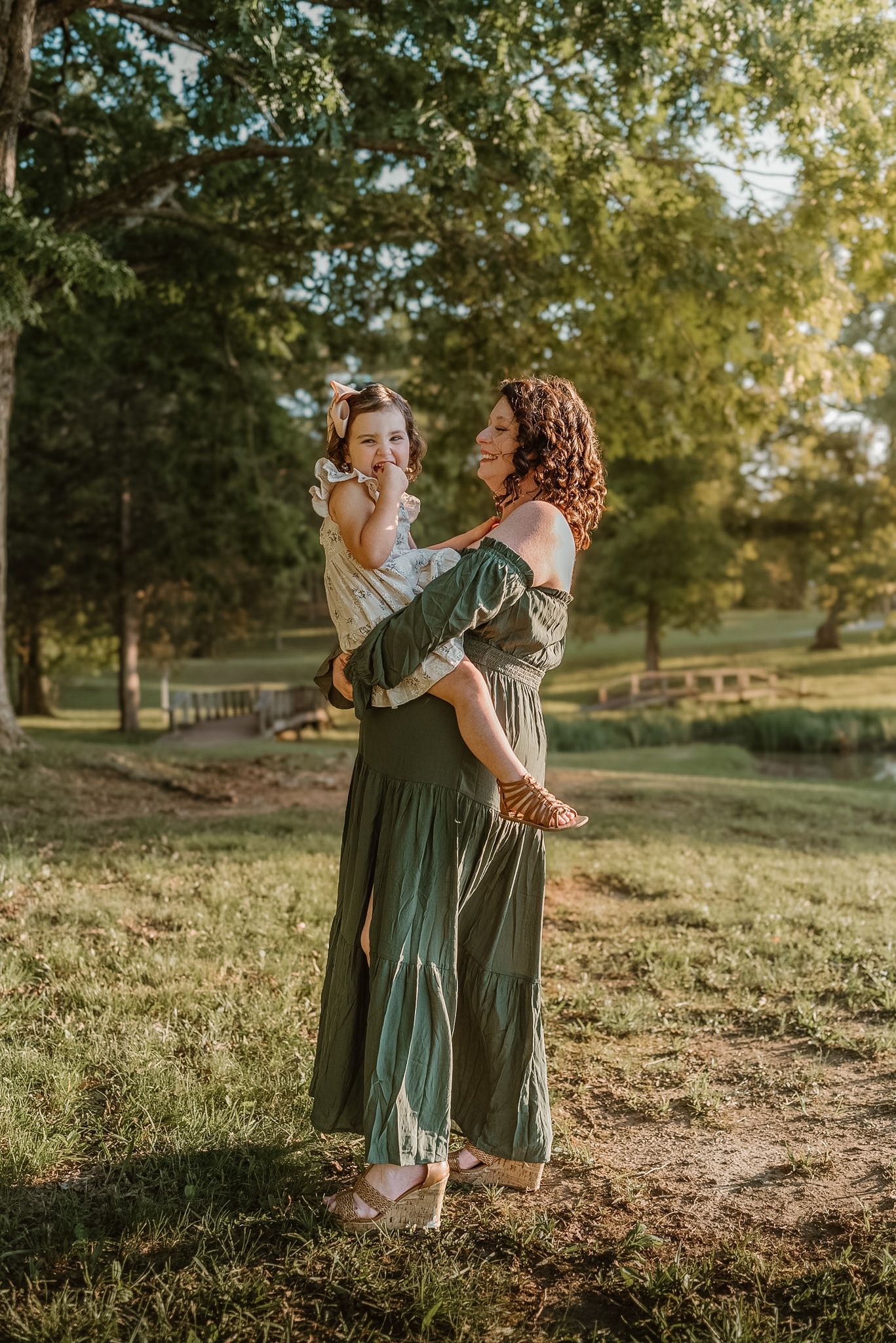 Mom holding toddler, both laughing with toddler looking at camera and mom looking at her in a field in Hixson, TN, warm golden light