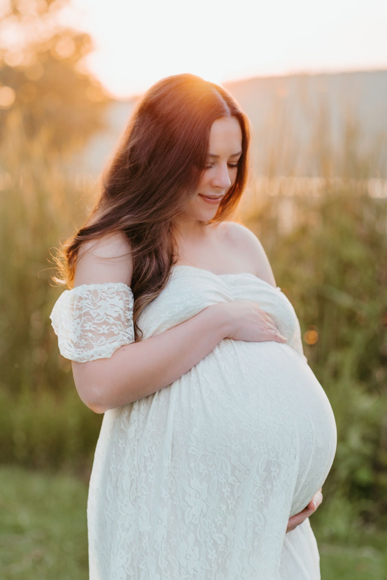 Expecting mom smiling down at baby bump with water and golden light behind her during maternity session in Jasper, TN