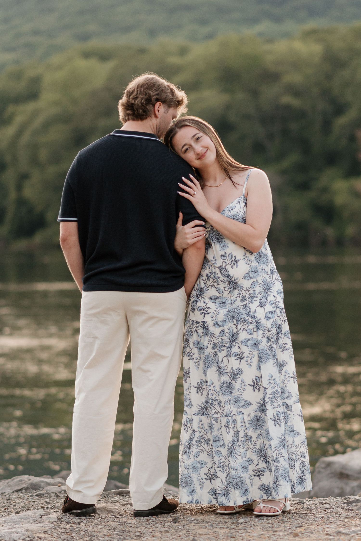 woman holding and resting her head on fiance's arm while he looks away towards river and raccoon mountain in evening engagement session