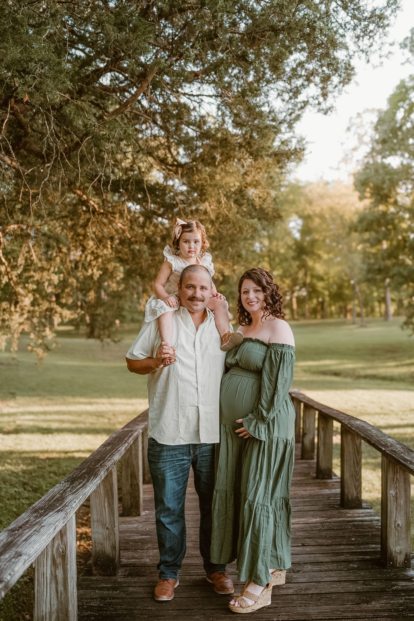 Posed family photo with toddler on dads shoulders, standing together on bridge and smiling at camera in a field in Hixson, Tennessee