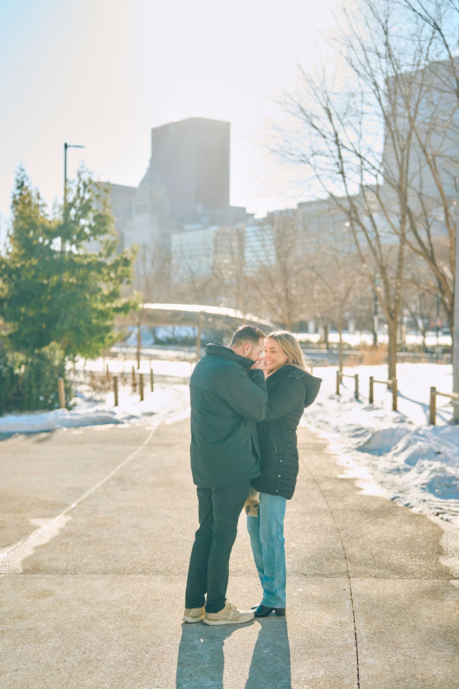 Two people embrace on a snowy winter path with city buildings visible in the background on a sunny day.