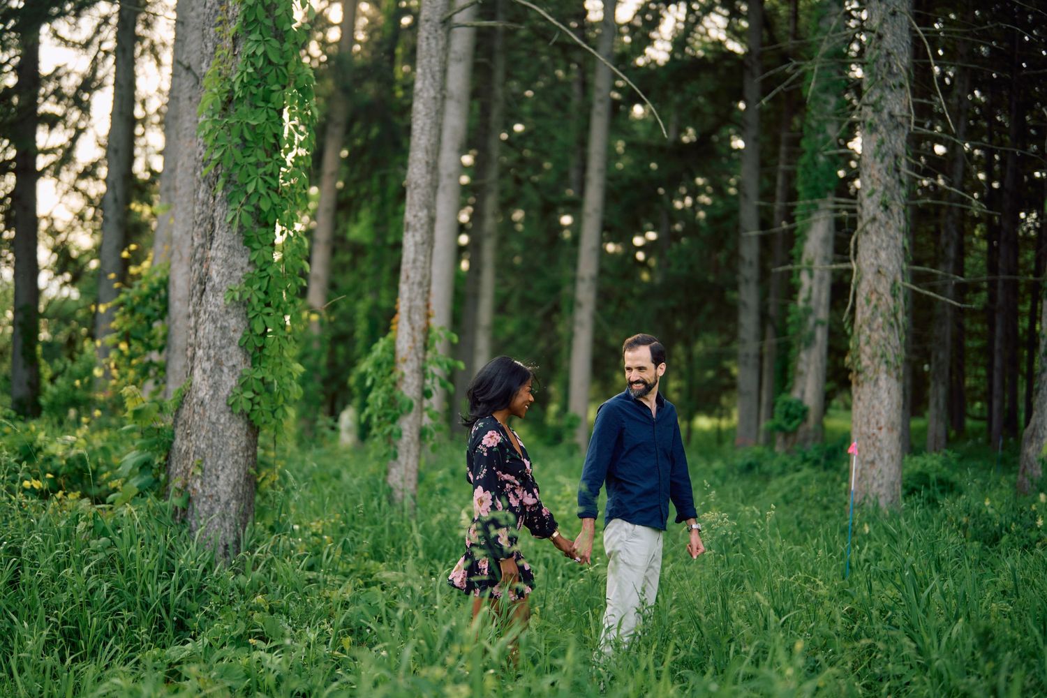 A couple shares a romantic moment while walking through a lush forest of tall pine trees on a sunny afternoon.