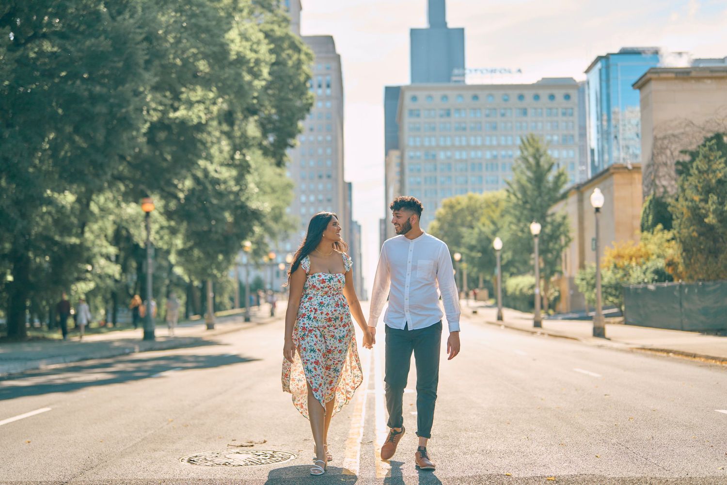 A couple walking together down an empty urban street with tall buildings and trees on a sunny summer day.