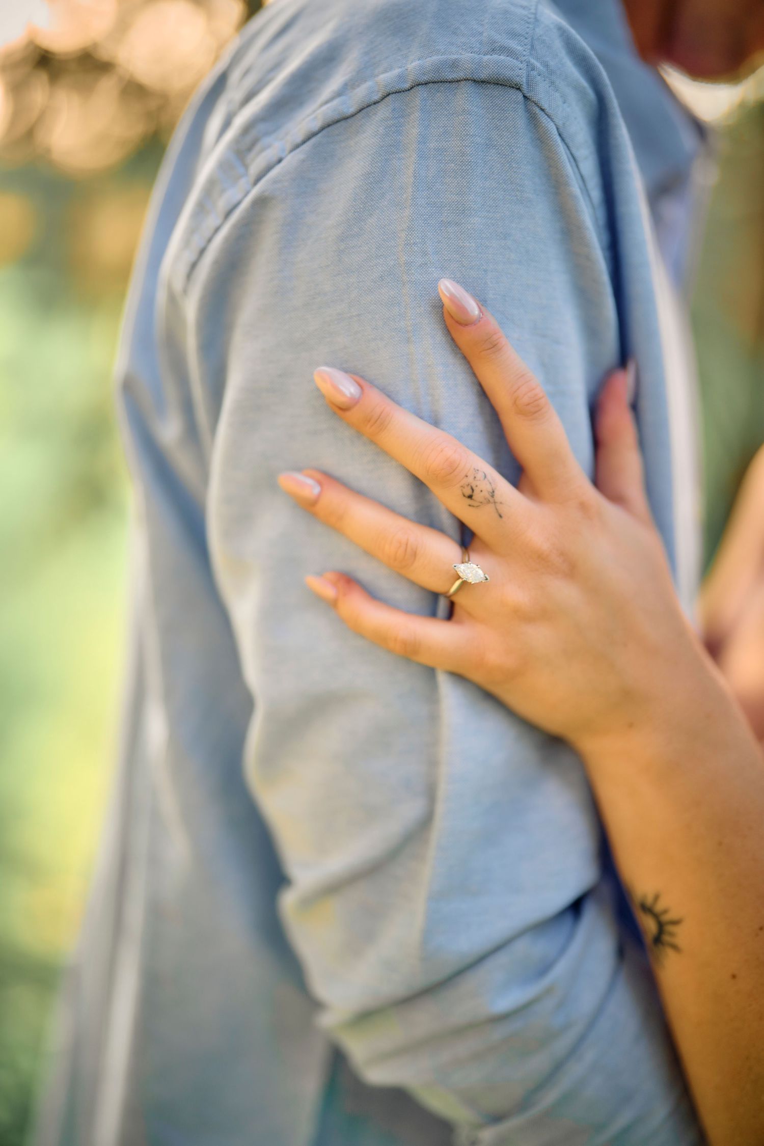 A delicate hand with neutral manicure and multiple rings rests on a light gray sleeve, showing a small wrist tattoo.