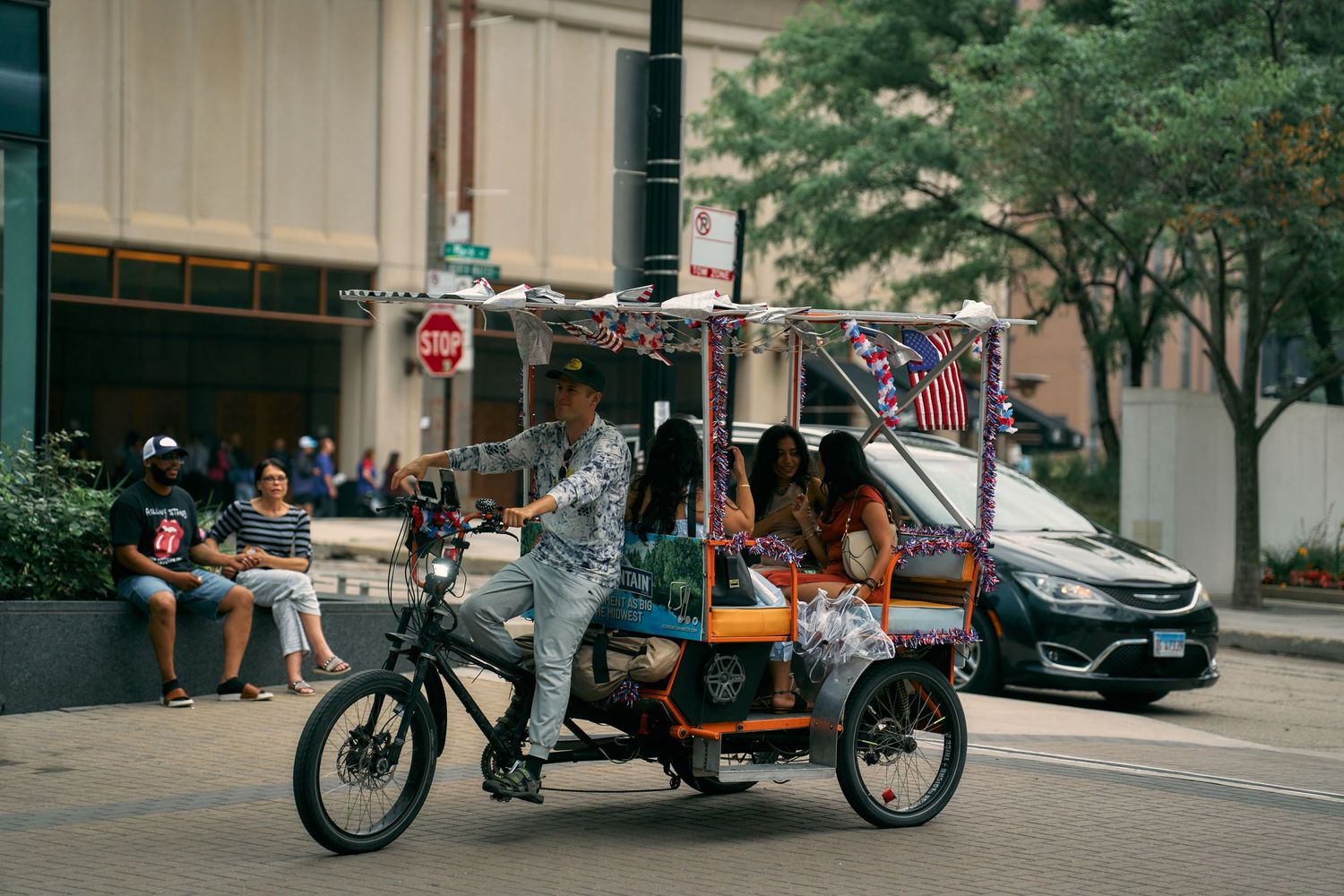 A pedicab driver operates their rickshaw on a city street with parked cars and pedestrians in the background.