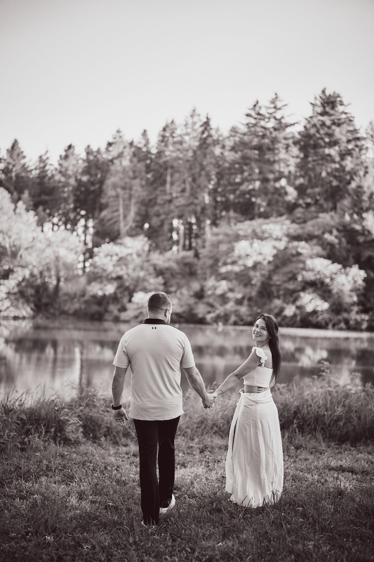 Couple walks hand in hand near a serene lake surrounded by pine trees in black and white photography.